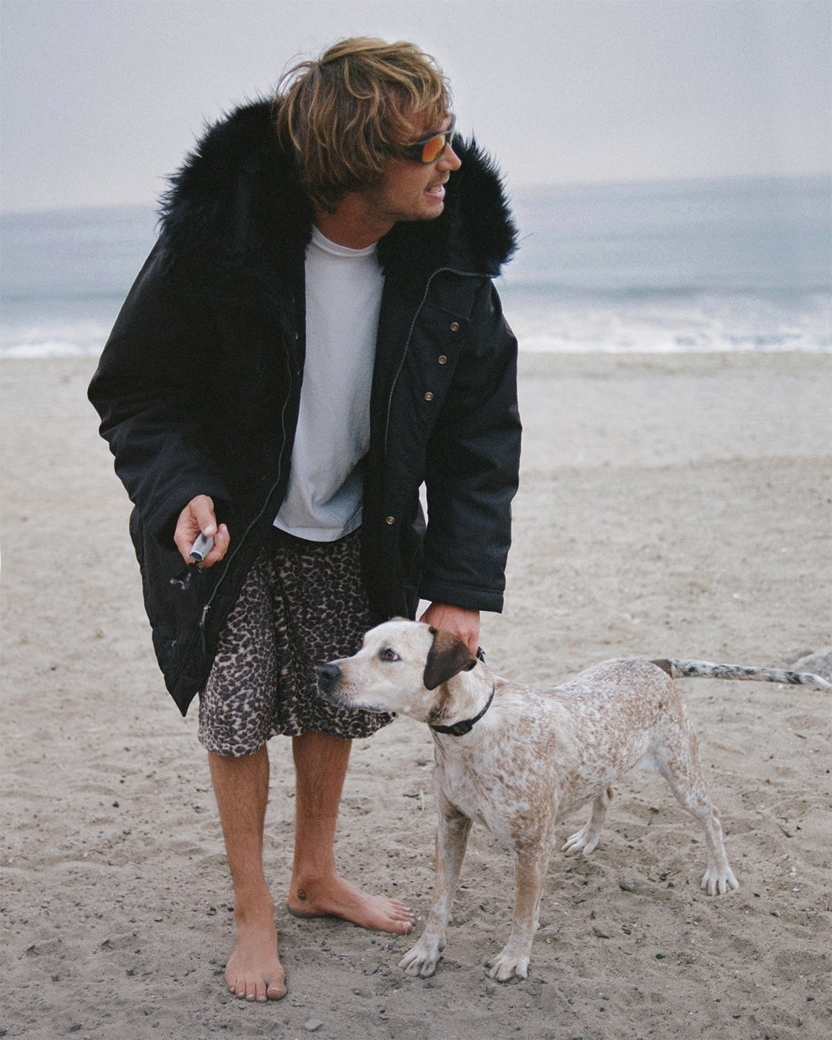 Person standing on a beach with a dog, wearing a black coat and sunglasses.