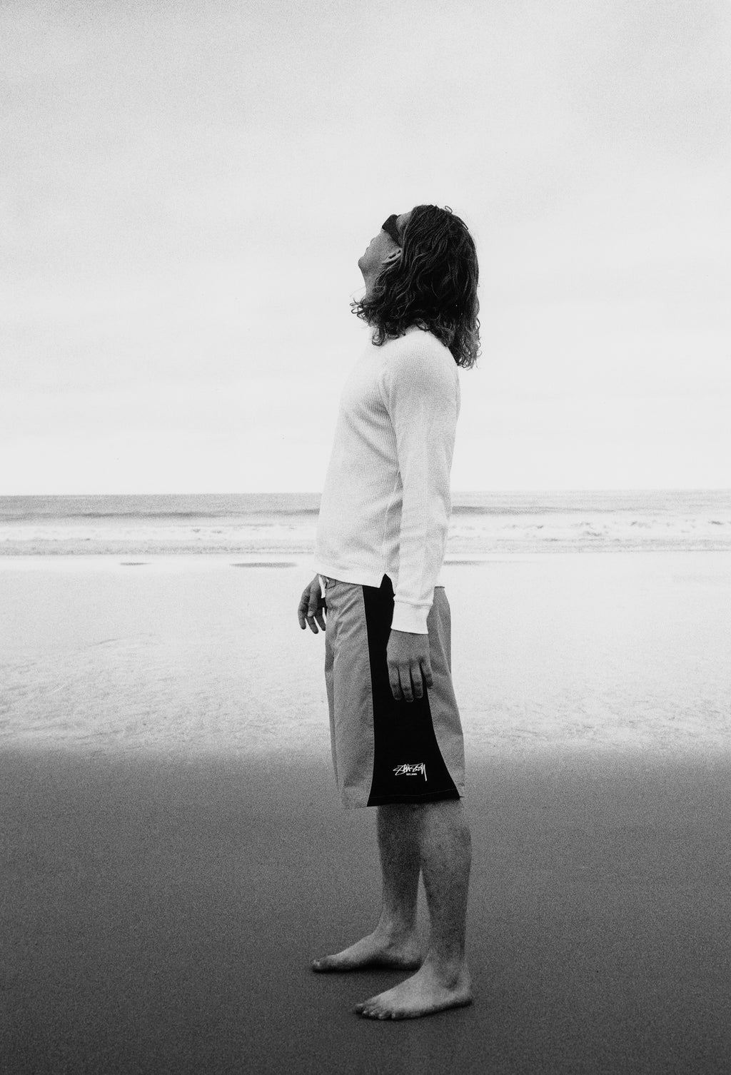 Model standing barefoot on a beach in the Pigment Dyed LS Thermal in White and the Curved Panel Board Short in Green, facing sideways with head tilted upward, with the ocean and horizon in the background.
