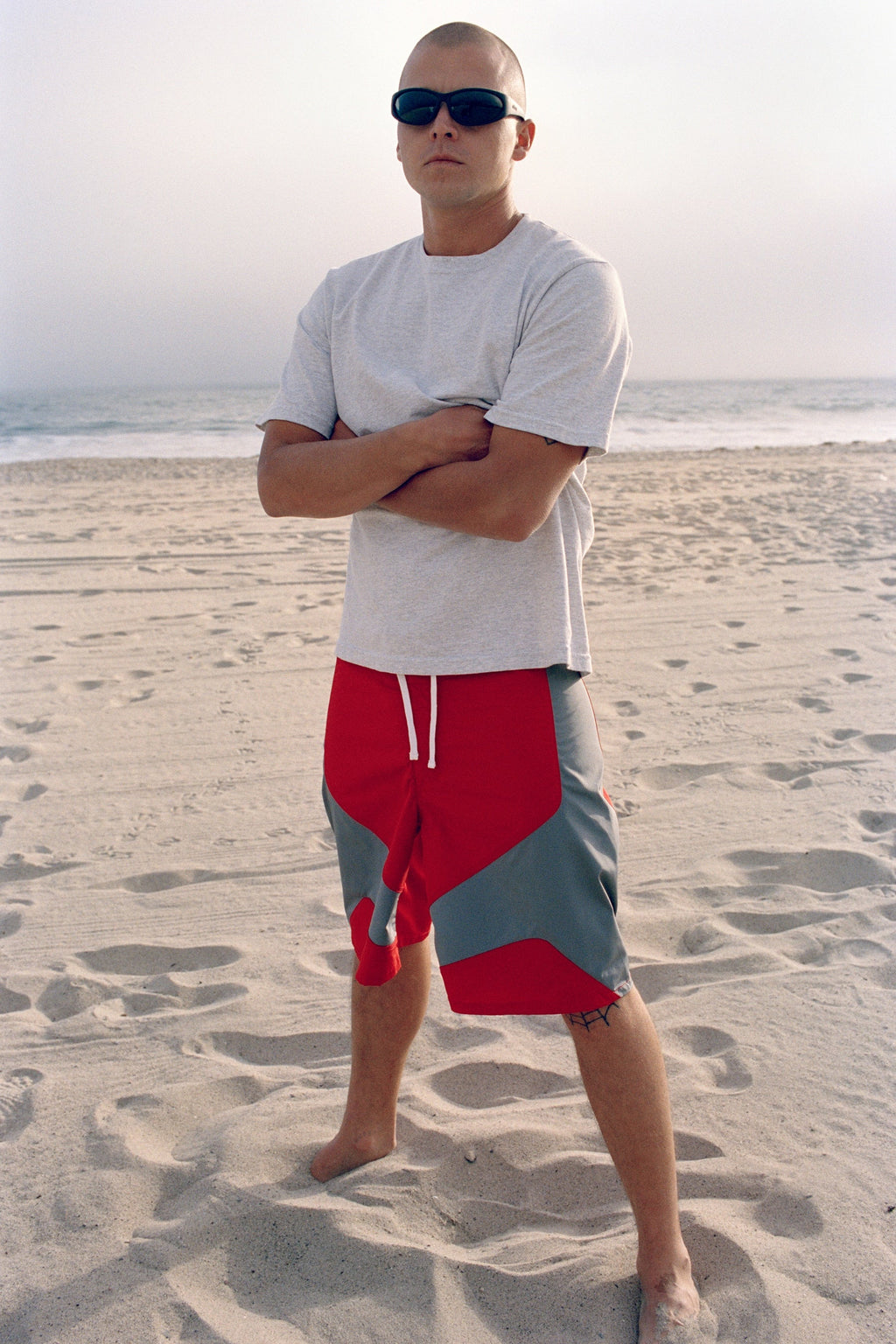 Model stands on a sandy beach wearing a grey t-shirt and Octane Board Short in Red.