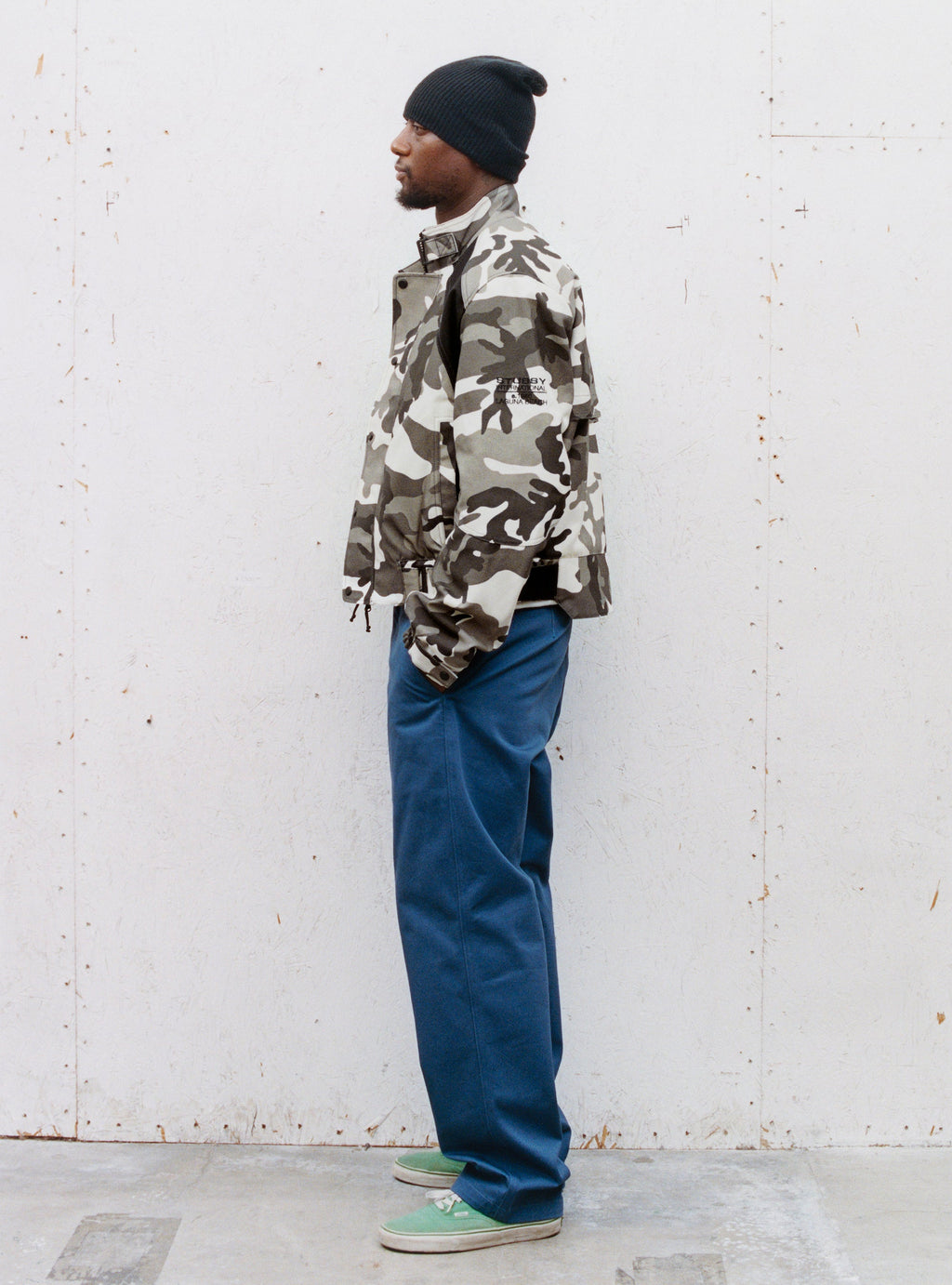 A man in profile stands against a white wall wearing the Touring Jacket in Snow Camo and Uniform Pant in Blue.