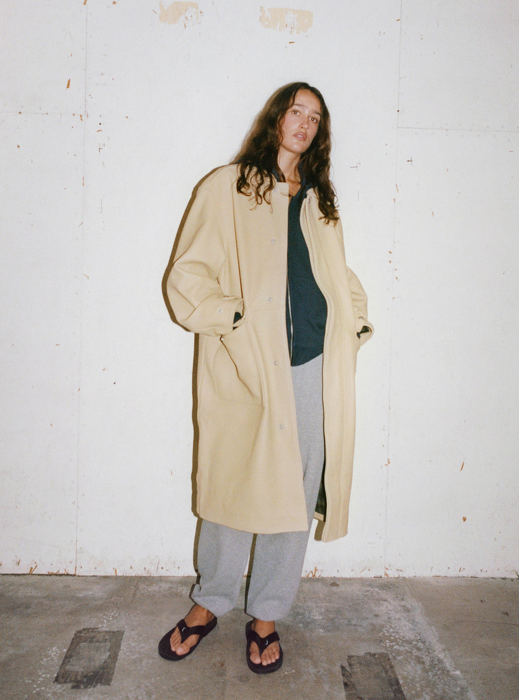 A model wearing the Leather Duster in Sand and Beach Slipper in Black stands in front of a white wall.