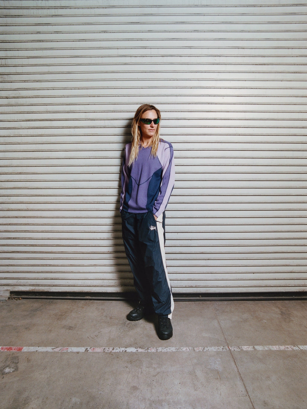 Full-length shot of a model with blonde hair standing against a closed metal shutter door, wearing dark athletic sunglasses, the Wave Knit Sweater in Lavender and Side Stripe Track Pant in Navy.