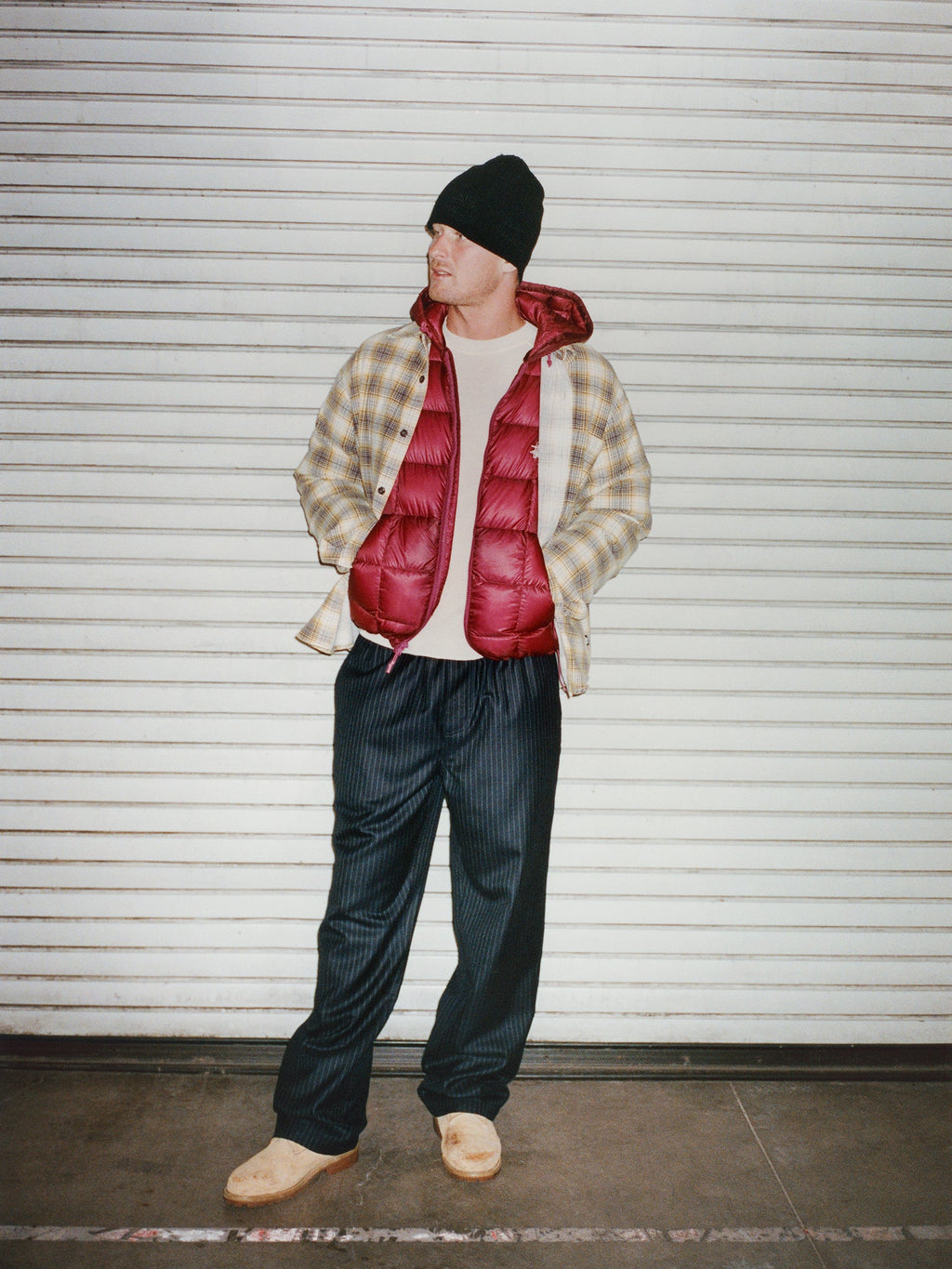 Full-length photo of a model wearing a black beanie, the Midweight Hooded Puffer in Fuchsia, Dax Printed Shirt in Gold and Striped Wool Beach Pant in Navy Pinstripe standing in front of a closed metal shutter door.
