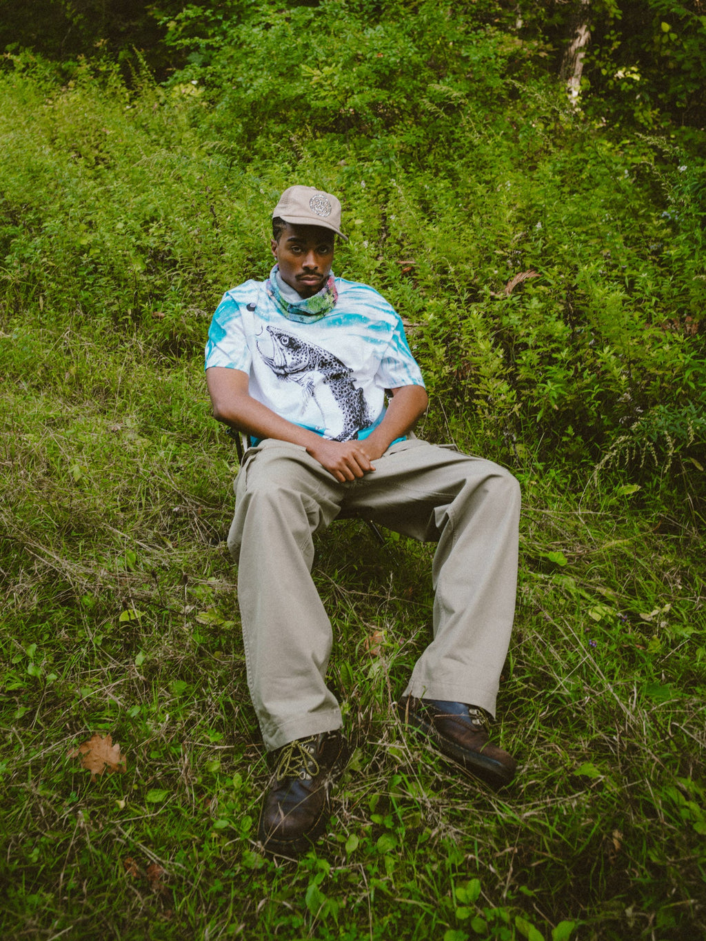 Photo of a model sitting on a chair surrounded by grass, wearing the Denim Tears Bass Tee and Denim Tears Cap