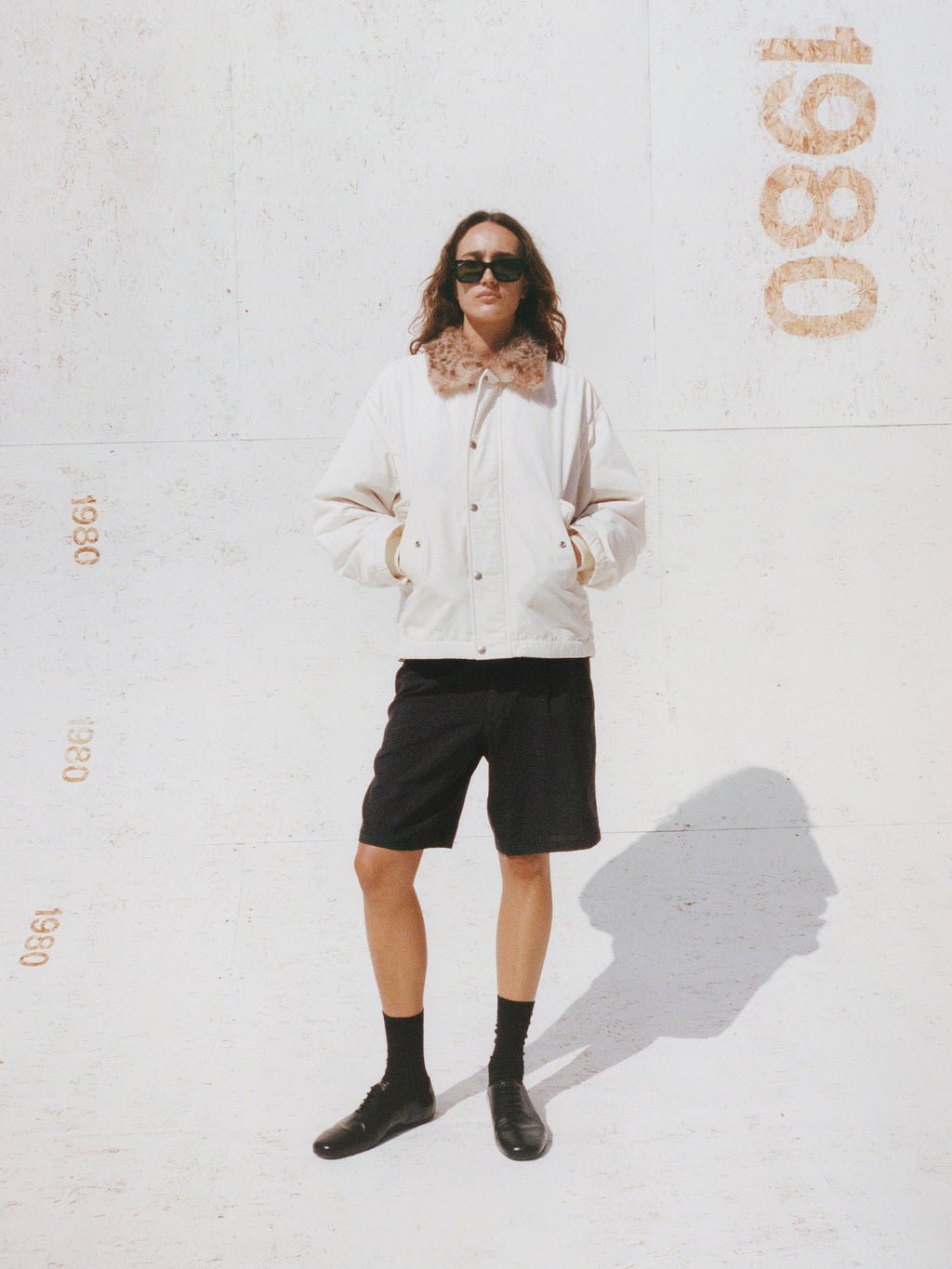 Model wears the Stüssy Convertible Deck Jacket in off-white with a faux-fur collar, styled with black shorts and black loafers.